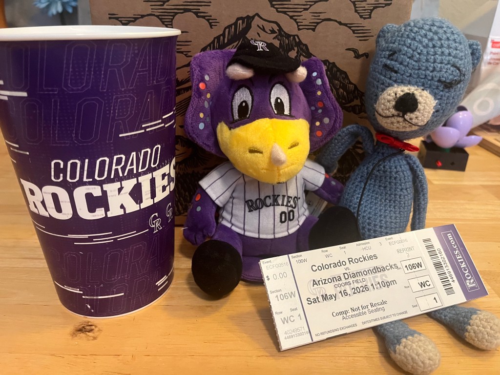 A Colorado Rockies cup next to a plush purple elephant mascot, a crocheted blue bear, and a baseball ticket for a game against the Arizona Diamondbacks on May 16, 2026.