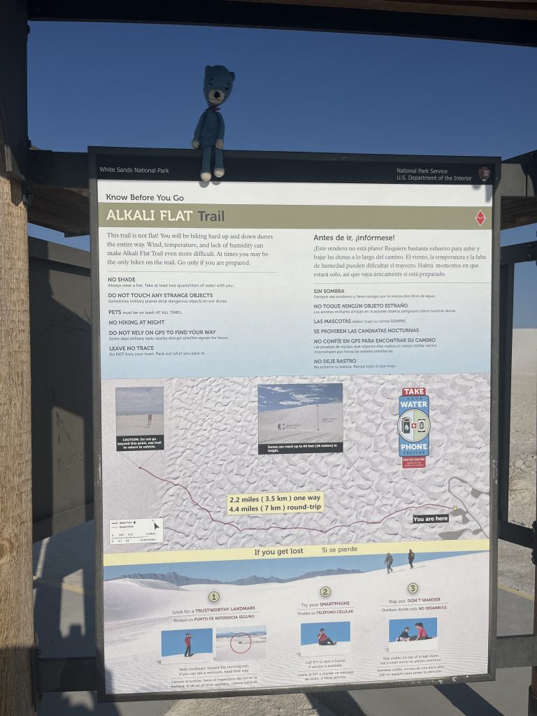Information sign for Alkali Flat Trail at White Sands National Park, featuring trail tips, safety guidelines, and distance markers. A blue stuffed bear is placed on top of the sign.