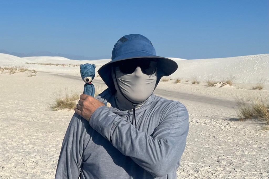 A person wearing a wide-brimmed hat and sunglasses stands in a sandy area, holding a small blue bear toy on their shoulder.