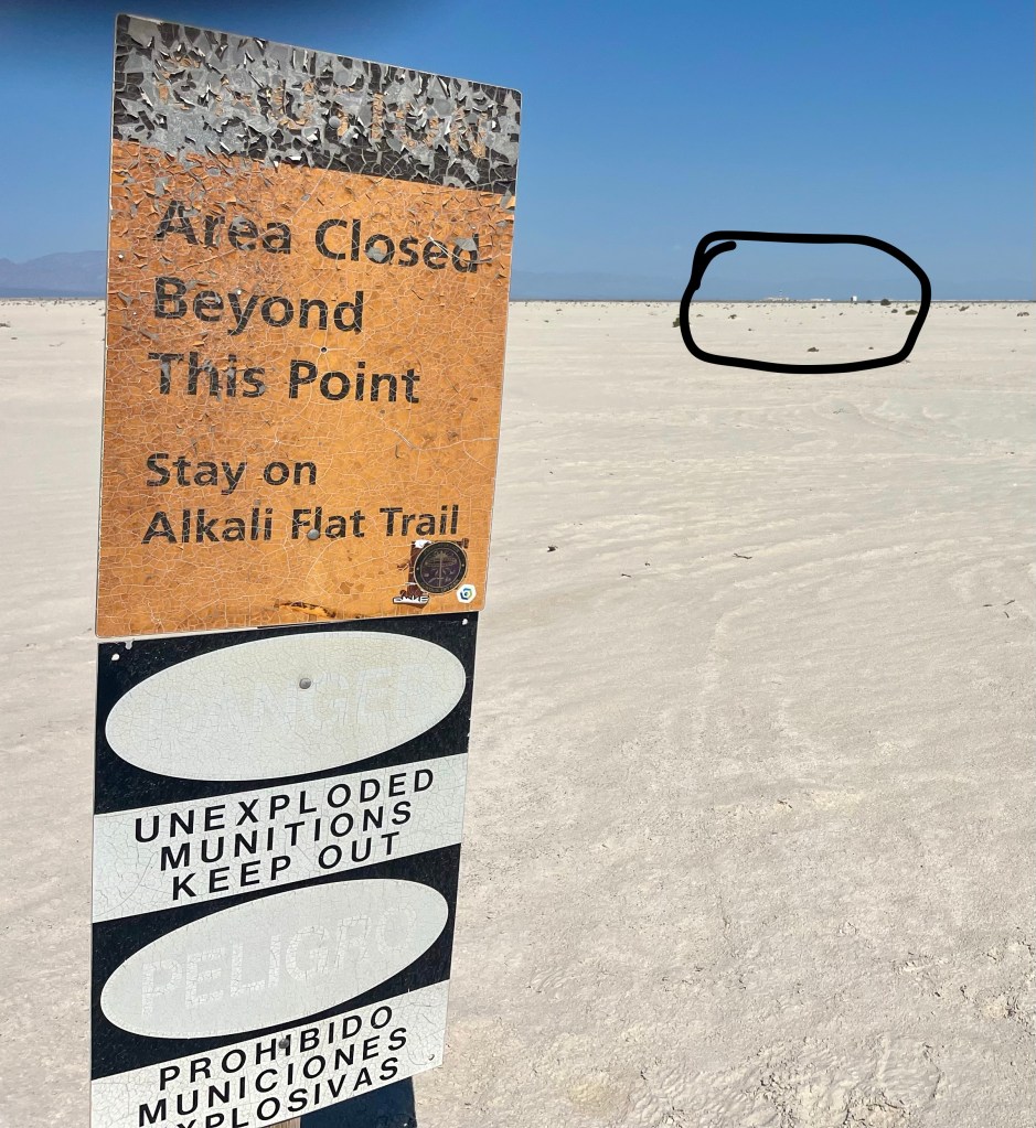 A sign indicating 'Area Closed Beyond This Point' at a sandy location, warning to stay on the Alkali Flat Trail and mentioning 'Unexploded Munitions, Keep Out'.